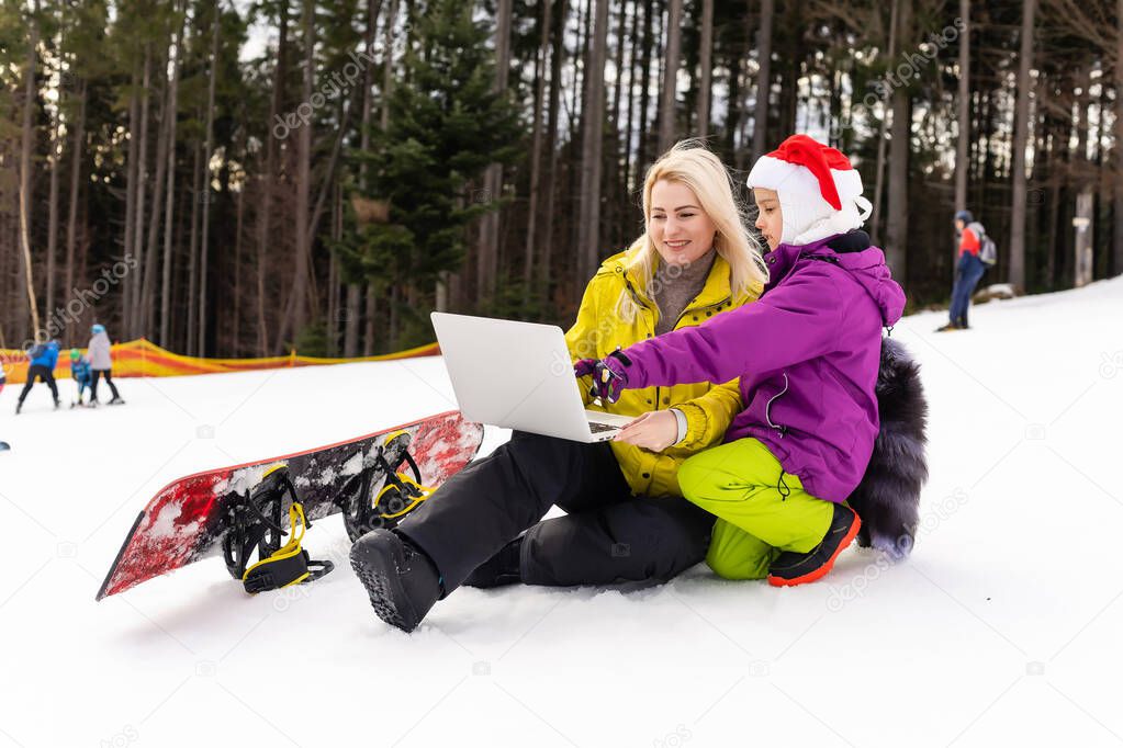 mother and daughter with a snowboard using laptop on snowy mountain