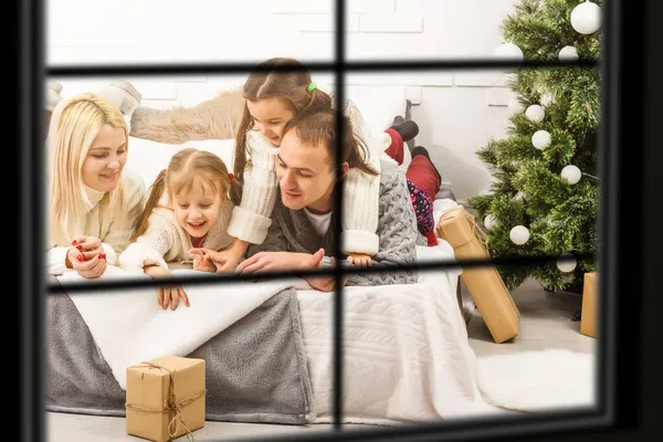 Giovane grande famiglia che celebra il Natale godendo la cena, vista dall'esterno attraverso una finestra in un soggiorno decorato con albero e luci a candela, genitori felici che mangiano con tre bambini . — Foto Stock