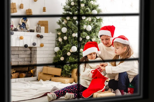 Joven gran familia celebrando la Navidad disfrutando de la cena, vista desde el exterior a través de una ventana a una sala de estar decorada con luces de árbol y velas, padres felices comiendo con tres niños . — Foto de Stock