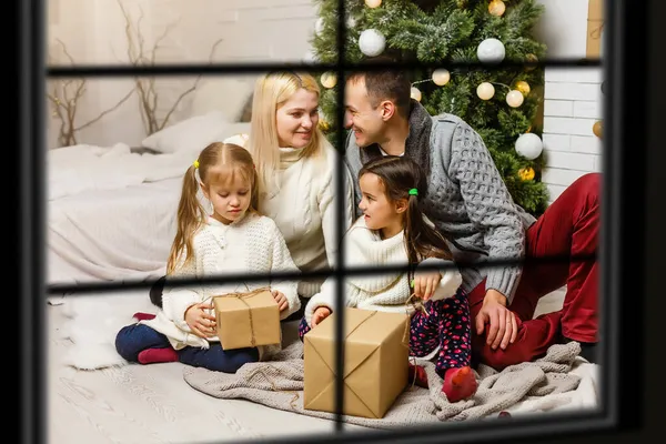 Joven gran familia celebrando la Navidad disfrutando de la cena, vista desde el exterior a través de una ventana a una sala de estar decorada con luces de árbol y velas, padres felices comiendo con tres niños . — Foto de Stock