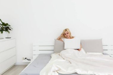 Yatağında dizüstü bilgisayarının arkasında oturan güzel, gülümseyen kadın.
