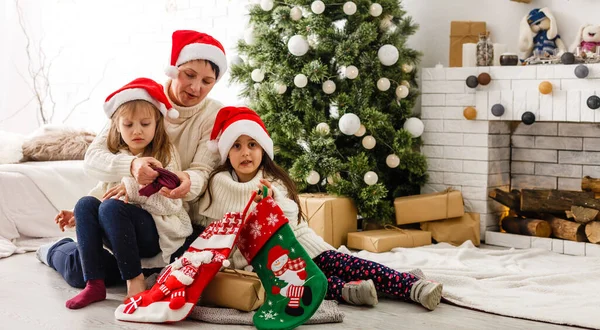 Wesołych Świąt i Wesołych Świąt. Wesoła mama i jej słodkie córki dziewczyny wymieniają się prezentami. Rodzic i dwójka małych dzieci bawią się razem w pobliżu choinki w domu. — Zdjęcie stockowe