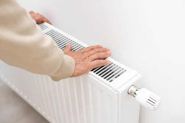 Een oudere man bevroor in zijn handen, een man verwarmt zijn handen in de buurt van een verwarming radiator in een appartement, een man bevriest in een appartement. — Stockfoto