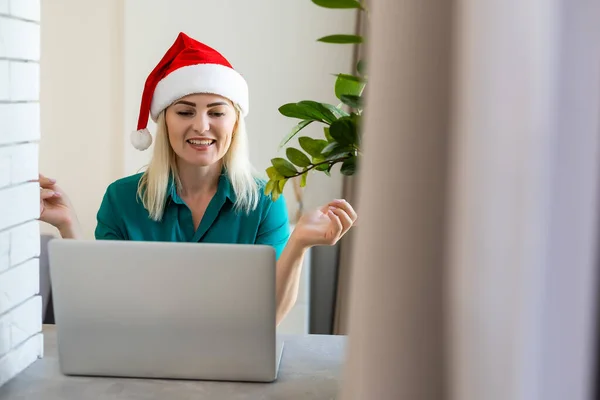 Mulher de chapéu vermelho Santa se comunica em uma videoconferência de Natal. A menina celebra o Natal em casa durante a pandemia. Comunicação com entes queridos através da Internet. Cumprimentos do ano novo online. — Fotografia de Stock