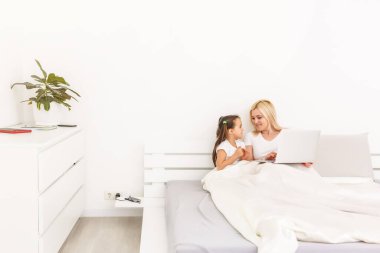 Anne ve kızı evde yatakta dizüstü kullanarak gülümseyen