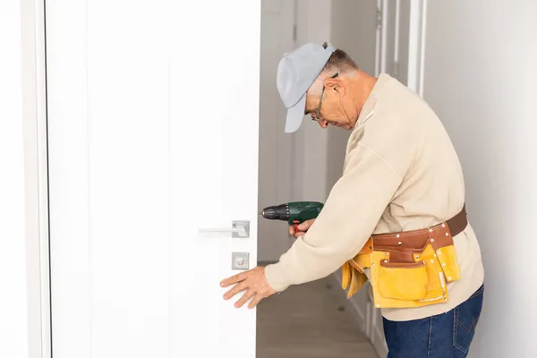 Attractive builder is installing lock in door. He is holding a screwdriver and kneeling. — Stock Photo, Image