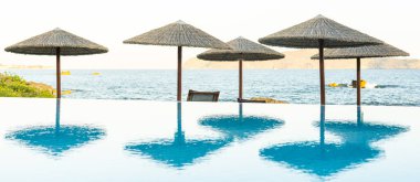 Güneş şemsiyesinin altındaki güverte sandalyeleri sonsuz bir havuz ve deniz arasında. Tepedeki alanı kopyala.