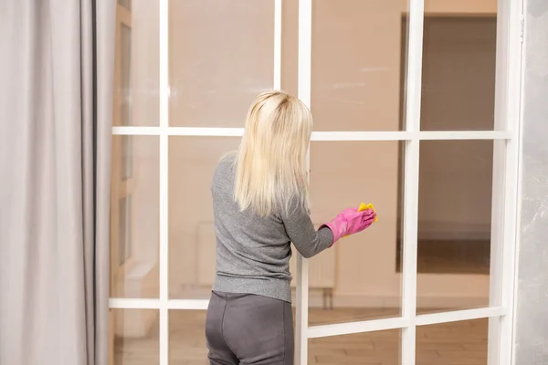 Visão traseira da mulher bonita em luvas de proteção segurando um spray e um pano enquanto limpa sua casa — Fotografia de Stock