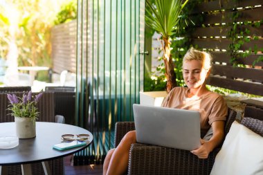 Genç bir bayan, halka açık kablosuz internet alanında dizüstü bilgisayarda çalışıyor, yazı yazıyor, insanlar arka planda geçip gidiyor.