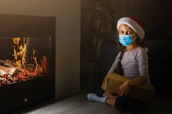 Noel ağacı arka planında koruyucu maske takan küçük bir kız. Cerrahi maskeli üzgün çocuk yüzü. Covid-19 pandemik konsepti boyunca Noel şenliği ve sosyal mesafeler — Stok fotoğraf