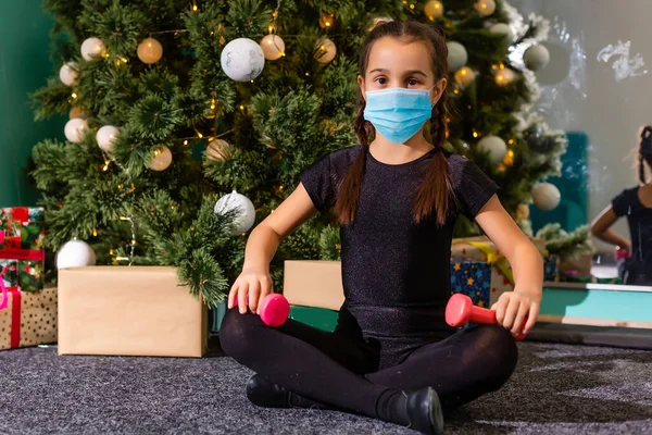 Noel ağacı arka planında koruyucu maske takan küçük bir kız. Cerrahi maskeli üzgün çocuk yüzü. Covid-19 pandemik konsepti boyunca Noel şenliği ve sosyal mesafeler — Stok fotoğraf