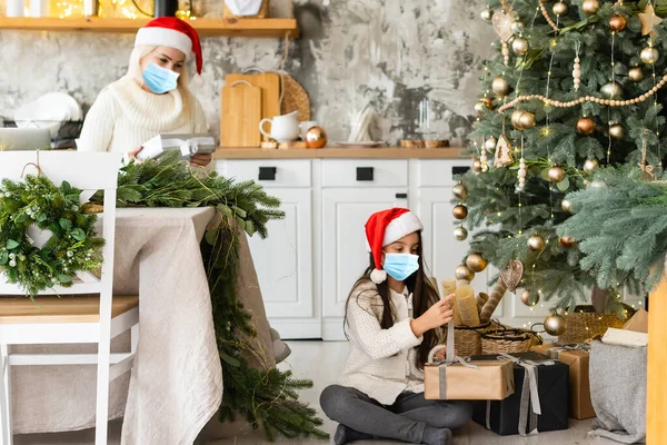 医療用マスクの美しい母親と娘は、屋内のクリスマスツリーの近くの家で楽しい時間を過ごすことができます。家族の幸福、休日、喜び、休暇、女性とのゲーム。新年の準備 — ストック写真