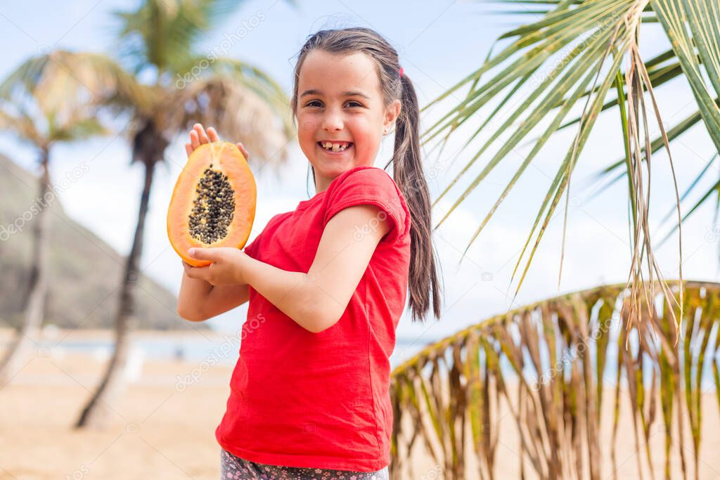 After playing on the sea black sand beach little cute girl picnics of natural vegetarian food - eating a orange slice of fresh ripe papaya, healthy children lifestyle in vacation on tropical island