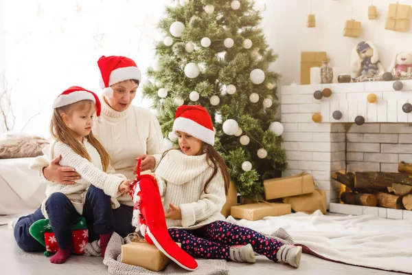 Junge Mutter und ihre beiden kleinen Töchter öffnen ein zauberhaftes Weihnachtsgeschenk am Weihnachtsbaum im gemütlichen Wohnzimmer im Winter — Stockfoto