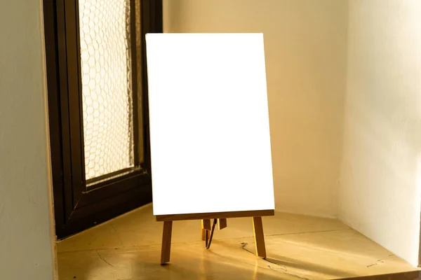 Cavalete de madeira com tela branca em branco fica perto da janela em um estúdio de arte, preparado para o trabalho do artista. atividades domésticas — Fotografia de Stock