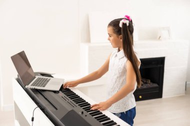 Evde eğitim gören küçük bir kız, okul öğretmeninin internet müziğine bağlı bilgisayardan piyano öğreniyor. Yeni normal yaşam tarzı ve eğitim, ev konseptinde öğrenim.