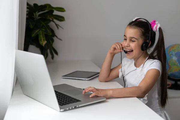 可爱的小女生在线聊天Skype笔记本电脑在线课上的视频电话课桌使用耳机向同学致意挥手隔离研究客厅室内. — 图库照片