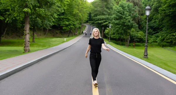 Joyful jovem senhora em roupas casuais andando ao longo da estrada de asfalto no campo, carona para passeio ao ar livre. Adorável mulher milenar viajando sozinha por autostop — Fotografia de Stock