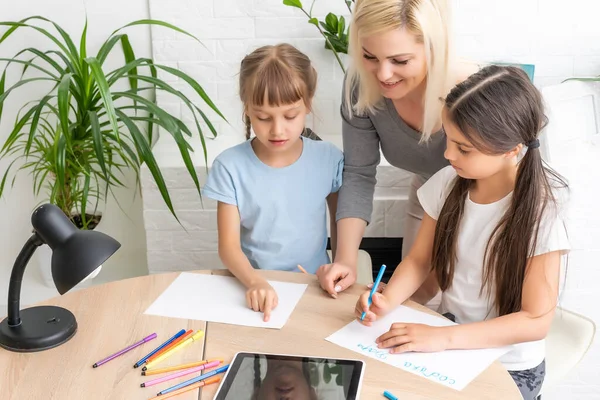 여교사나 양어머니가 귀여운 초등 학교 여학생 이 책상에 앉아 숙제하는 것을 도와 주고 있습니다. 어머니와 자녀 가집에서 공부하면서 공책에서 쓰는 법을 배우는 모습 — 스톡 사진