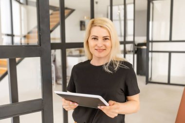 Pencerenin kenarında oturan ve evde dijital tablet kullanan genç bir kadının fotoğrafı..