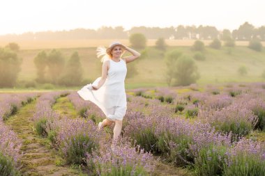 Beyaz elbiseli, genç ve güzel bir kadın lavanta tarlasında neşeyle koşuyor. Hasır şapkalı.