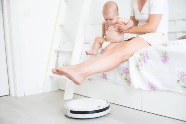 Genç anne yeni yürümeye başlayan oğluyla kanepede oturuyor ve robot elektrik süpürgesinin ev işi yapmasını izliyor..