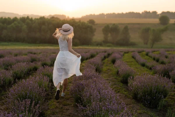 Piękna młoda zdrowa kobieta w białej sukience biegnąca radośnie przez pole lawendy, słomkowy kapelusz — Zdjęcie stockowe