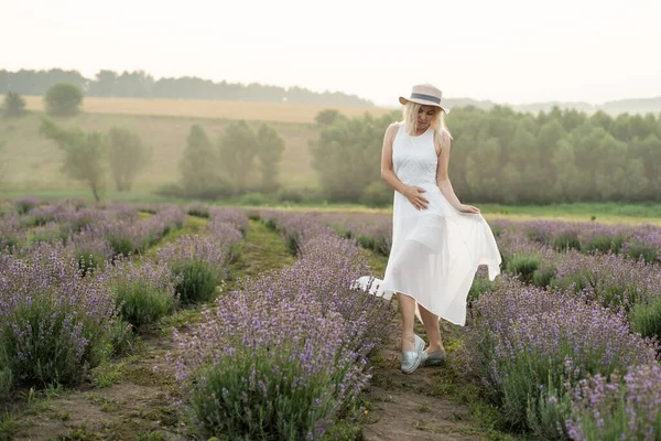 Mulher saudável jovem bonita com um vestido branco correndo alegremente através de um campo de lavanda, chapéu de palha — Fotografia de Stock