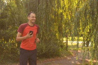 Tişörtlü yakışıklı bir adam parkta antrenman yapıyor ve kulaklıkla müzik dinliyor.