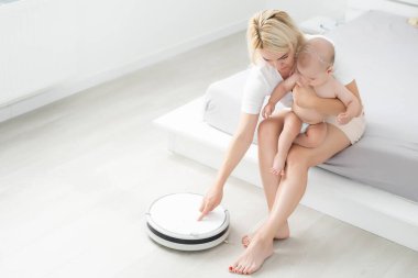 Genç anne yeni yürümeye başlayan oğluyla kanepede oturuyor ve robot elektrik süpürgesinin ev işi yapmasını izliyor..