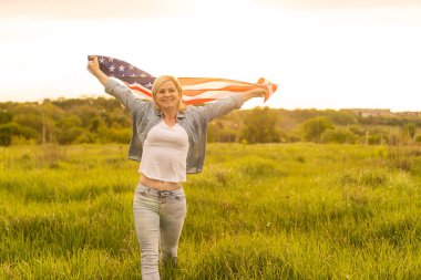 Tarlada rüzgarda Amerikan bayrağı tutan çekici bir kadın. Mavi gökyüzüne karşı yaz manzarası. Yatay yönelim.