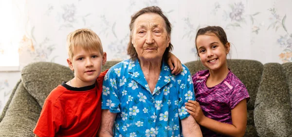 子供との曾祖母。家族だ。小学生の男の子と幼児の女の子。室内で幸せな年配の女性と孫たち. — ストック写真