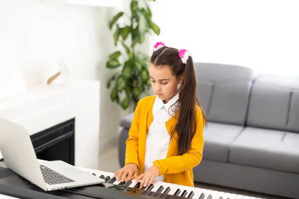 어린 소녀가 컴퓨터를 통해 학교 선생님에 의해 인터넷 음악 교실에 연결되는 피아노를 배우고 있습니다. 새로운 생활 방식 과 교육, 가정에서 공부하는 학생. — 스톡 사진