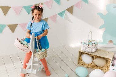 Paskalya 'ya hazırlanıyorum. Paskalya yumurtası tutan ve arka planda süslemelerle gülümseyen sevimli küçük kız.