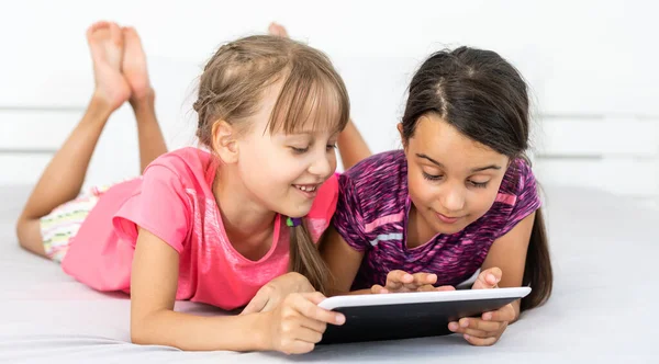 Dos niñas lindas están utilizando una tableta digital y sonriendo mientras están acostados en la cama en la habitación de los niños — Foto de Stock