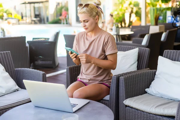 Une photo vue de côté de la femme utilisant un ordinateur portable pour le e-shopping tout en étant assis dans un hall d'hôtel moderne. Une femme blonde portant une robe regarde l'écran — Photo