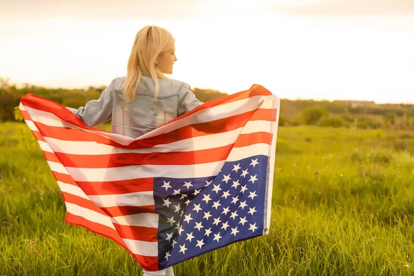 Tarlada rüzgarda Amerikan bayrağı tutan çekici bir kadın. Mavi gökyüzüne karşı yaz manzarası. Yatay yönelim. — Stok fotoğraf