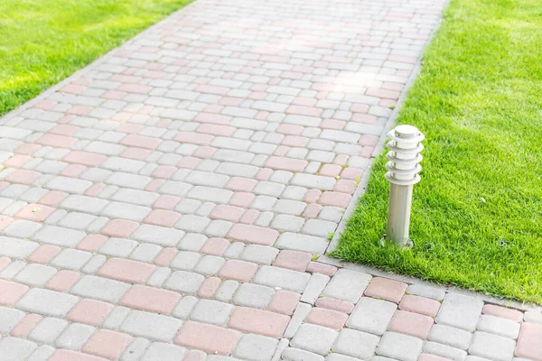 Ground iron lantern on a winding curved pedestrian walkway of red tiles for walks in the park with a green lawn and plants on a sunny summer day, nobody. — Stock Photo, Image