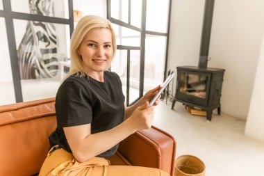 Kadın tablet üzerinde akıllı ev kontrolü uygulaması kullanıyor. Arka planda oturma odasının içi..