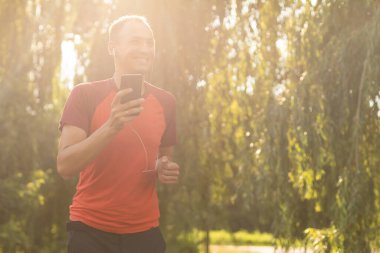 Tişörtlü yakışıklı bir adam parkta antrenman yapıyor ve kulaklıkla müzik dinliyor.