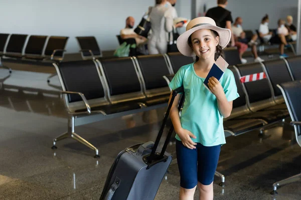 Turismo, vacaciones, infancia y concepto de transporte - niña sonriente con bolsa de viaje, boleto y pasaporte sobre el fondo del aeropuerto. — Foto de Stock