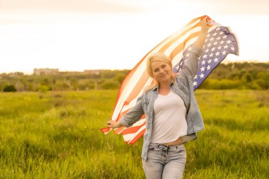 Tarlada rüzgarda Amerikan bayrağı tutan çekici bir kadın. Mavi gökyüzüne karşı yaz manzarası. Yatay yönelim.