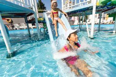 Yazın iki kız açık havuza su sıçratır. Mutlu çocuklar, kız kardeş oynuyor, halka açık havuzda güneşli havanın tadını çıkarıyor.