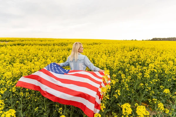 Mujer adulta sosteniendo bandera americana con poste, estrellas y rayas en un campo de colza amarillo. Bandera de EE.UU. ondeando en el viento — Foto de Stock