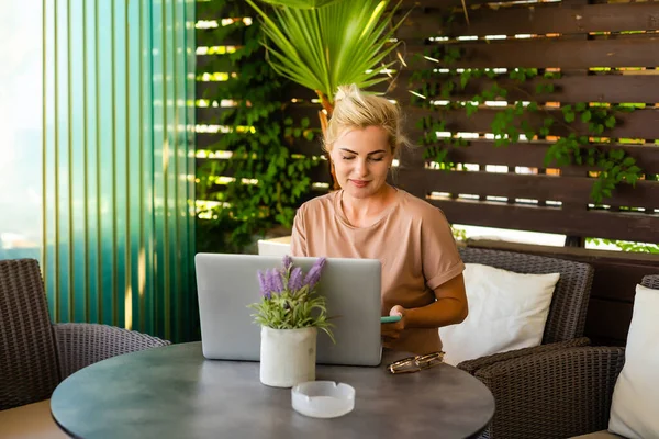 Μια πλαϊνή άποψη φωτογραφία της γυναίκας που χρησιμοποιεί φορητό υπολογιστή για e-shopping, ενώ κάθεται σε ένα σύγχρονο λόμπι του ξενοδοχείου. Μια ξανθιά γυναίκα που φοράει φόρεμα κοιτάζει την οθόνη. — Φωτογραφία Αρχείου