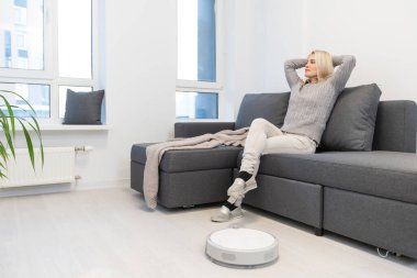 Kadın kanepede dinlenirken robot süpürge odayı temizliyor. Uzaktan kumandayla vakumu kontrol eden kadın.