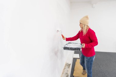 Kapatın. Yeni evde tek başına resim yapan oldukça neşeli beyaz bir kadının yan görüntüsü. Güzel bir kadın evi yeniliyor. Duvarı paten fırçasıyla boyuyor. Daireyi yeniden tasarlıyor.