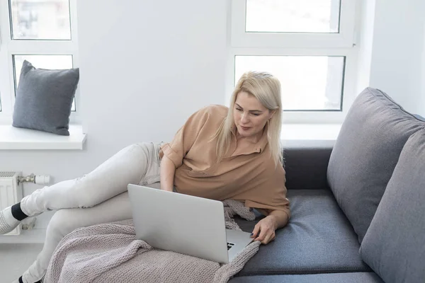Sorrindo mulher feliz sentado no sofá e usando laptop. O conceito de trabalho remoto ou estudo durante a quarentena — Fotografia de Stock