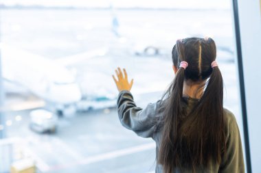 Küçük kız havaalanı geçiş salonundaki uçağına binmek için bekliyor ve kalkış kapısının yanındaki uçak parkına bakıyor. Çocukla birlikte hava yoluyla seyahat etmek