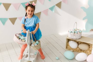 Paskalya 'ya hazırlanıyorum. Paskalya yumurtası tutan ve arka planda süslemelerle gülümseyen sevimli küçük kız.